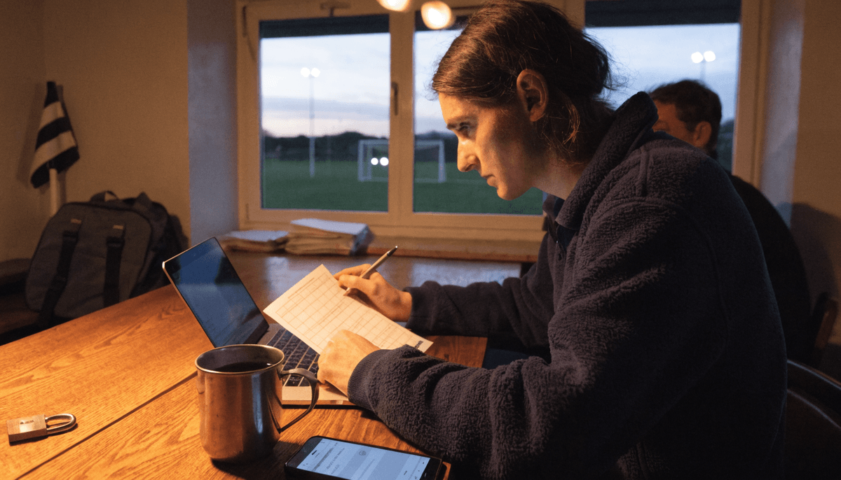 Volunteer club secretary in a clubhouse committee room at dusk working through a printed checklist beside a laptop, phone and padlock, with a floodlit pitch through the window