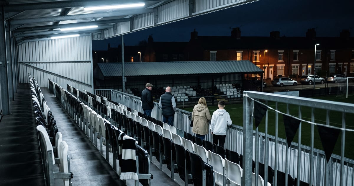 A high corner view at night inside a compact English non-league football ground, looking down long diagonal rows of empty black and white tip-up seats with a black-and-white supporters' scarf draped over one seat back in the foreground; four distinct spectators — a man in a flat cap, a stocky man in a black gilet, a woman in a beige raincoat and a teenager in a white hoodie — stand widely spaced along the front rail seen from behind, with a low covered stand opposite, black bunting on a railing, and red-brick terraced houses, lit windows, modern parked cars and street lighting beyond under a dark blue night sky; no faces, no text, no crest.