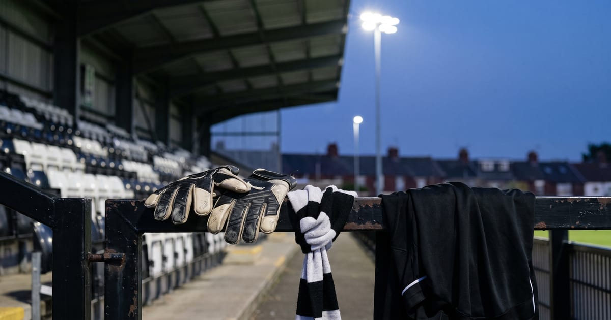 A blue-hour dusk still-life at a compact black-and-white English National League ground in Tyneside: a worn pair of black and white goalkeeper gloves rests on a chipped black metal crush barrier beside a knotted black-and-white supporters' scarf and a black training top, with rows of empty black and white seats under a corrugated covered stand and red-brick terraced rooftops soft-focus behind under distant floodlight glow; no people, no text, no crest.
