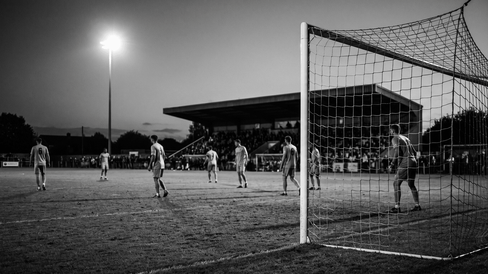 Non-league football ground under evening floodlights during a cup tie, small main stand and worn pitch, players in plain kits